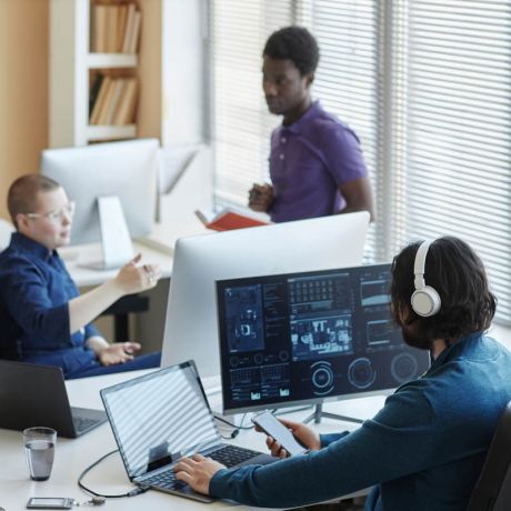 Young worker of cyber security center sitting in front of computers with graphic data and using smartphone while his colleagues having discussion